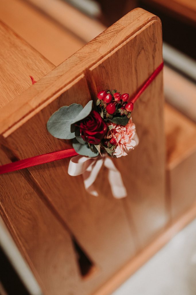 bukiecik z czerwonych i beżowych kwiatów ze wstążkami jako dekoracja ławek w kościele do ślubu