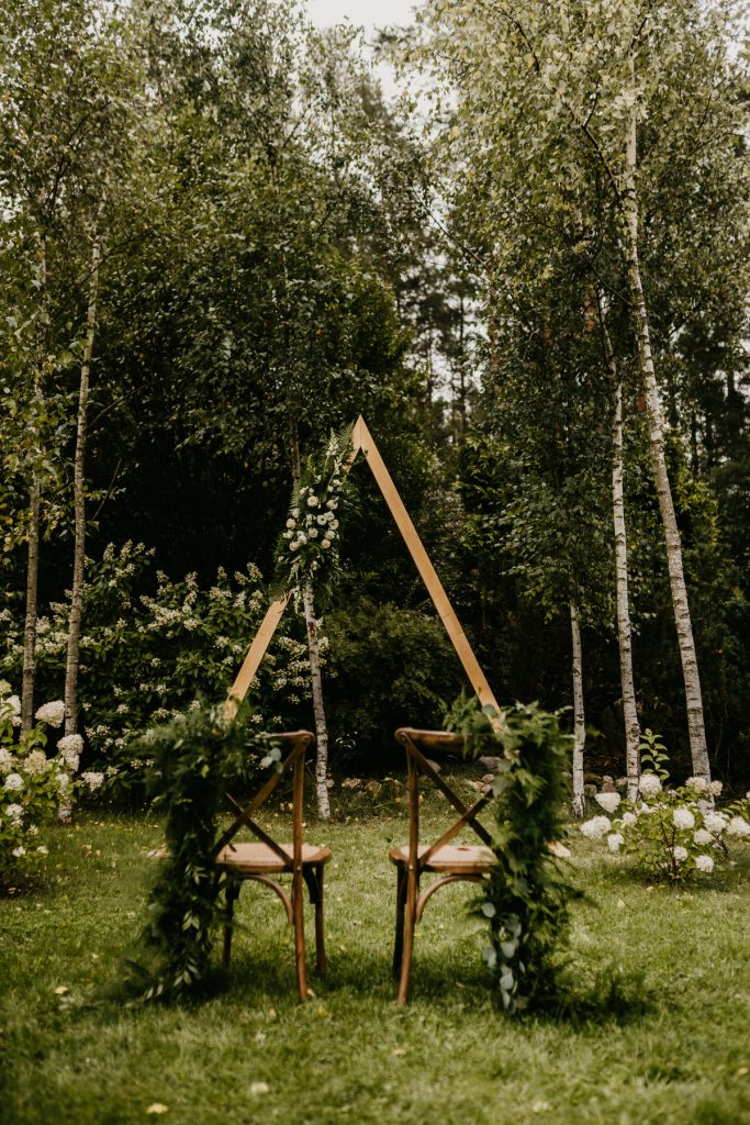 trójkąt drewniany z kwiatami i krzesła boho z girlandami do dekoracji ślubu w plenerze