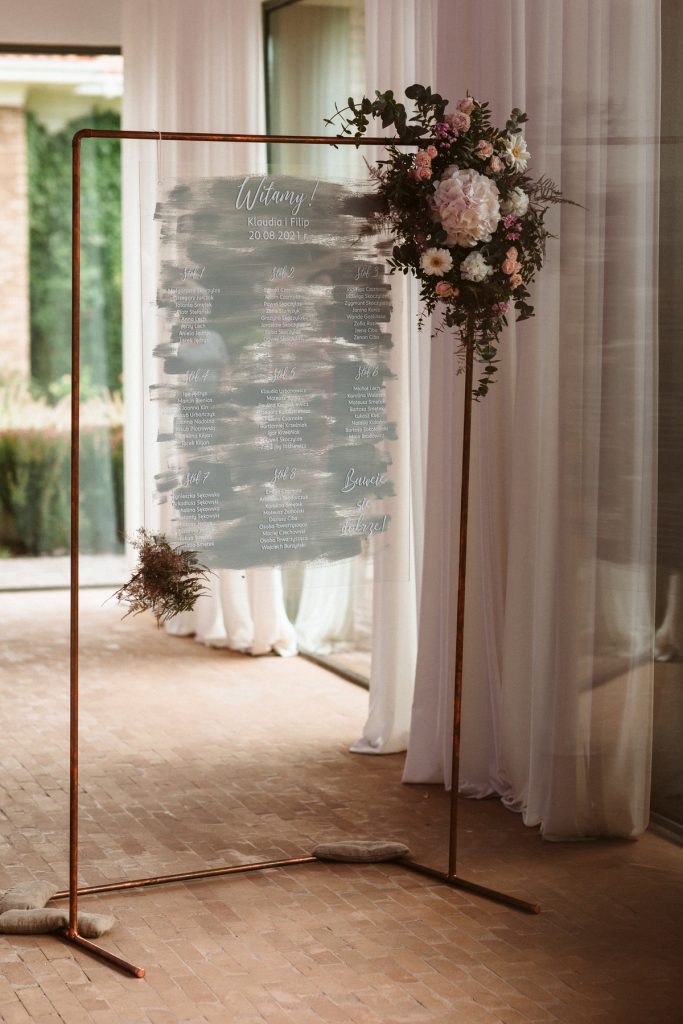 stojak miedziany jako plan usadzenia gości na tablicy z pleksi z dekoracją kwiatową