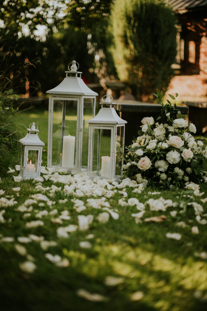 świece w białych lampionach z płatkami róż na trawie prowadzącej do ślubu